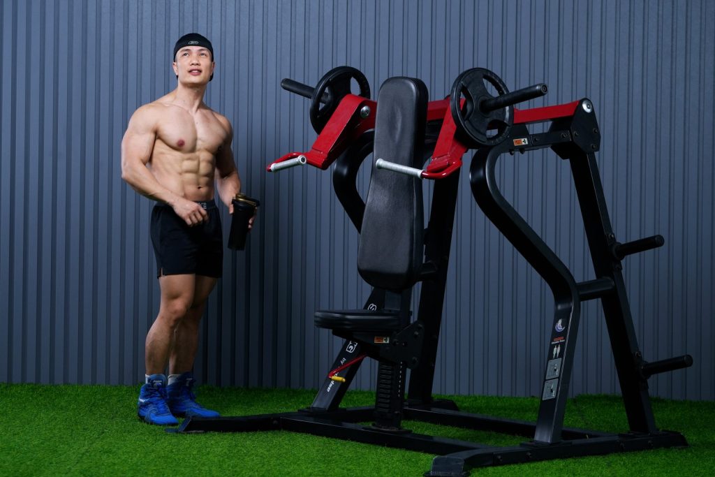 Huấn luyện viên Nguyễn Bảo Bằng cởi trần khoe cơ bụng sáu múi, tay cầm bình lắc, đứng cạnh máy đẩy vai trong phòng tập