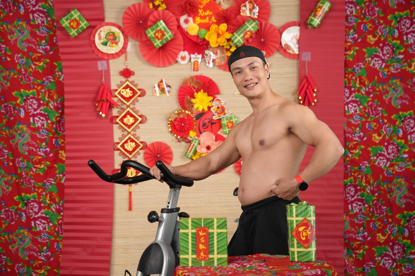 Nguyễn Bảo Bằng đứng cạnh xe đạp tập, tay véo lớp mỡ bụng, nền phông trang trí Tết có bánh chưng và câu đối