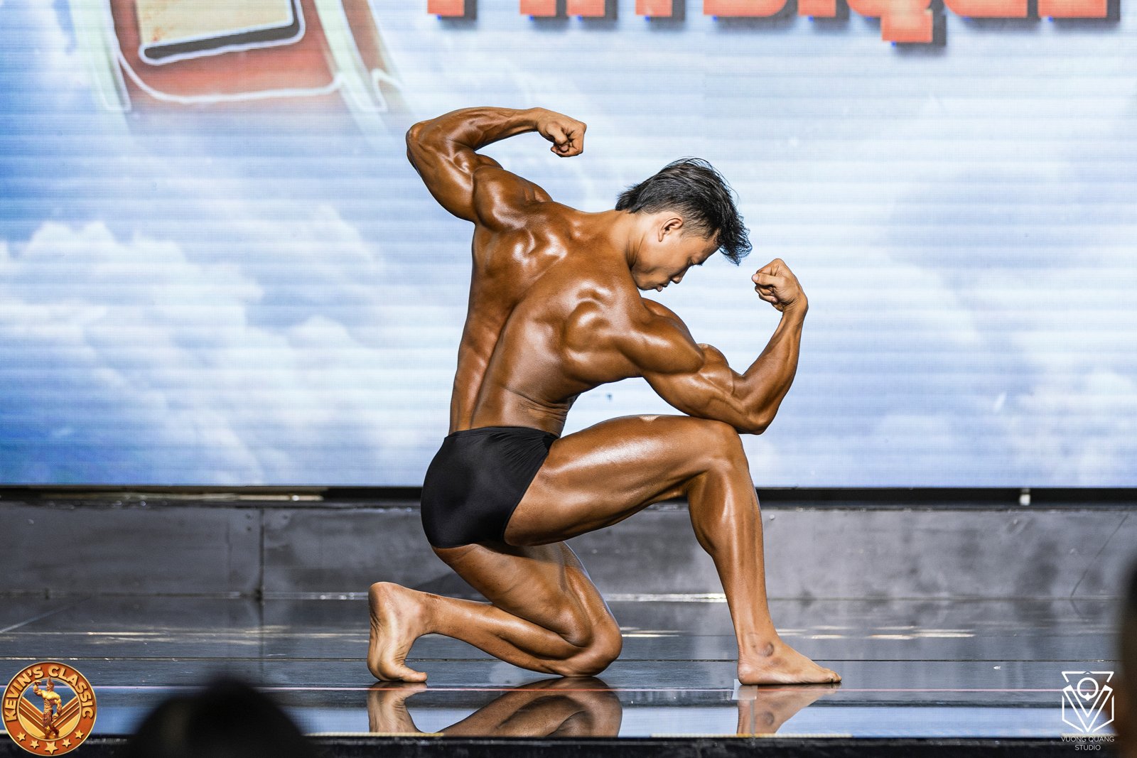 Vận động viên Classic Physique quỳ một gối khoe cơ tay và lưng