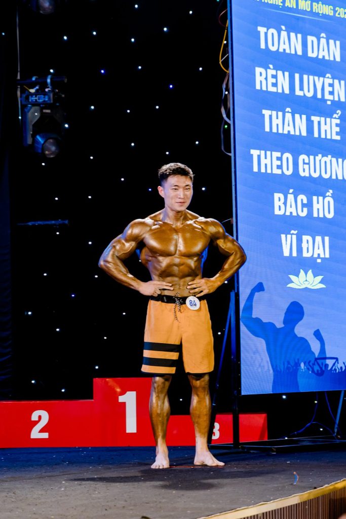 Vận động viên men's physique đứng cạnh bục 1-2-3, phía sau là băng-rôn Toàn dân rèn luyện thân thể theo gương Bác Hồ vĩ đại