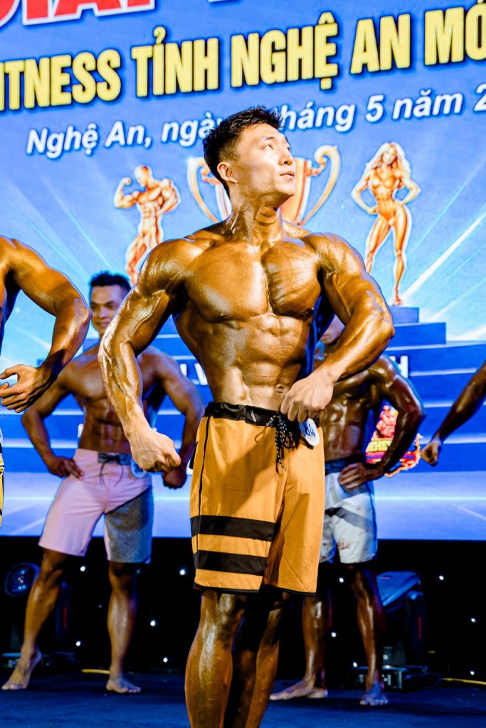 Vận động viên men's physique tạo dáng mặt trước, phông và hình cúp phía sau tại giải thể hình Nghệ An 2026