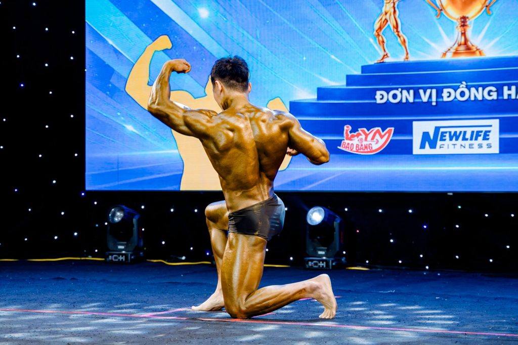 Vận động viên thể hình quỳ gồng lưng bên dòng Đơn vị đồng hành và logo Newlife Fitness tại giải thể hình Nghệ An 2026