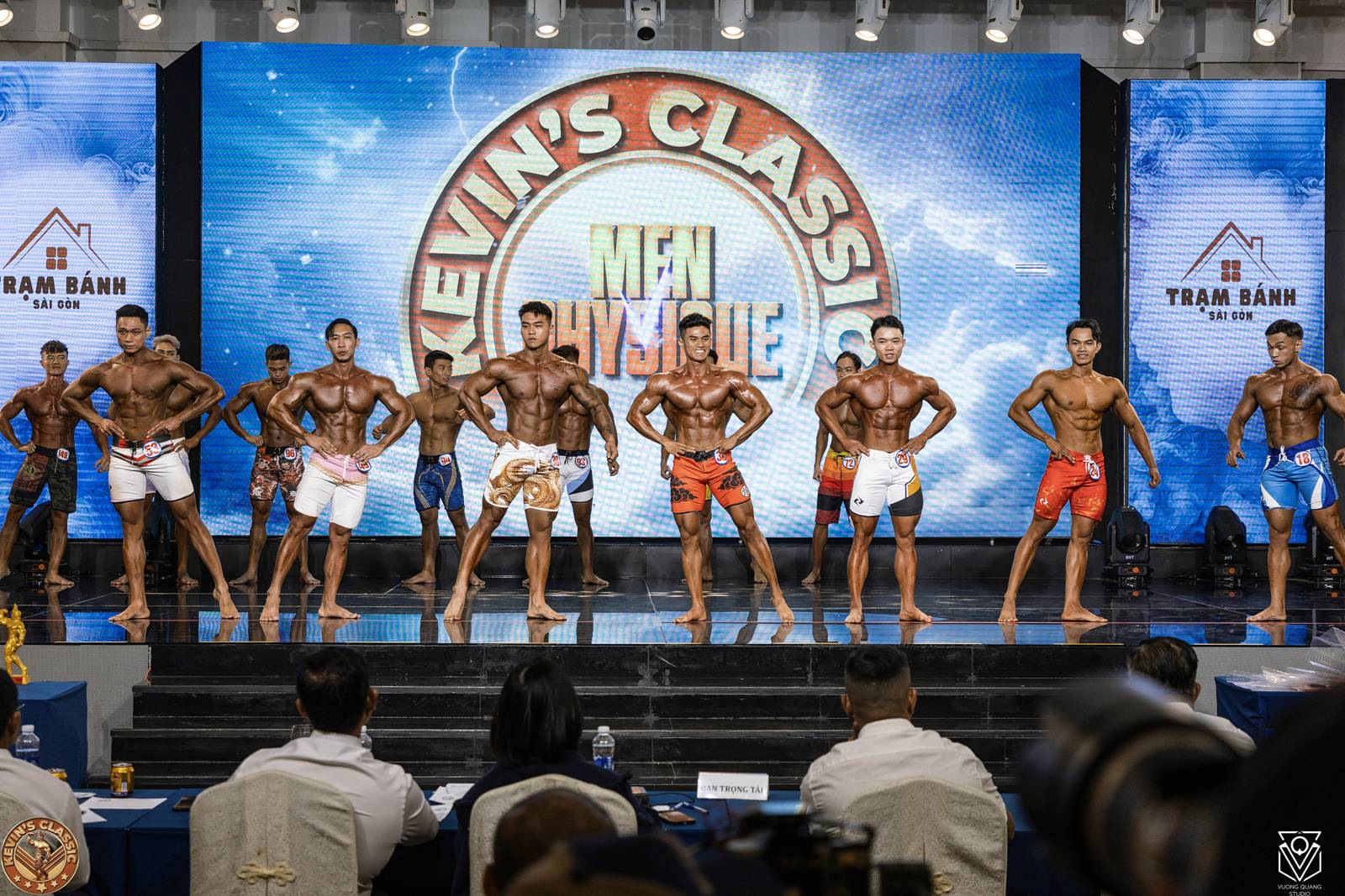 Hàng vận động viên Men Physique đứng trước bàn trọng tài