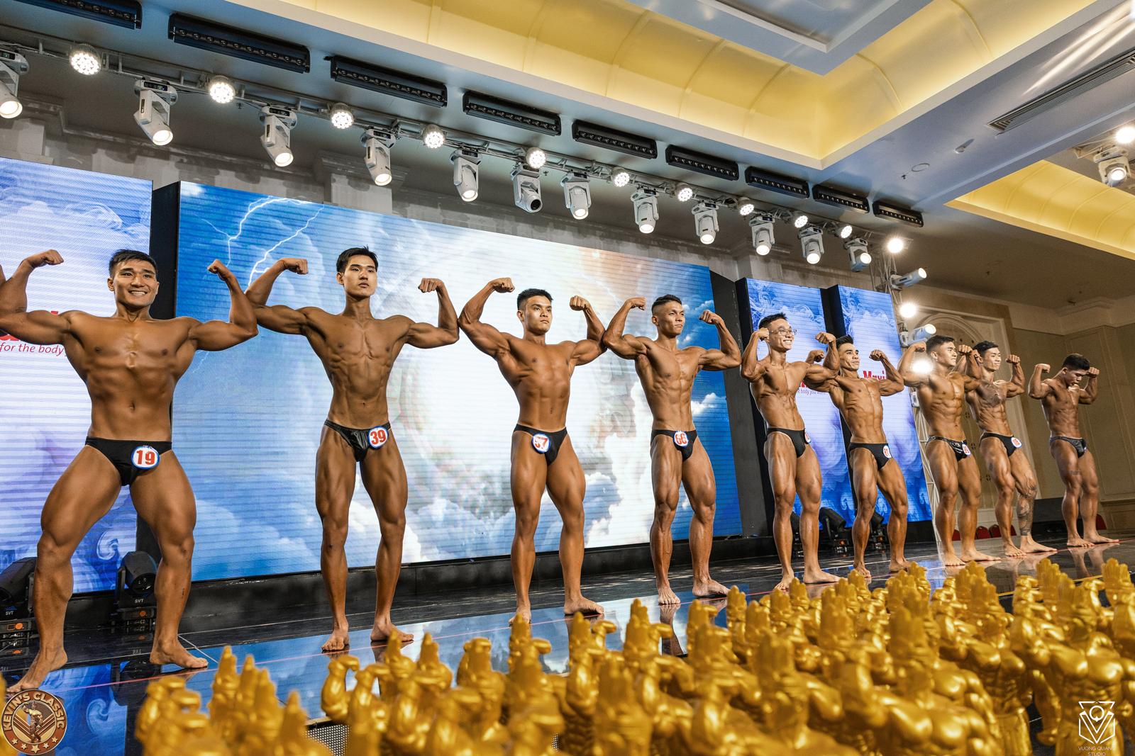 Hàng vận động viên gồng cơ trong một lượt so dáng, cúp vàng ở tiền cảnh