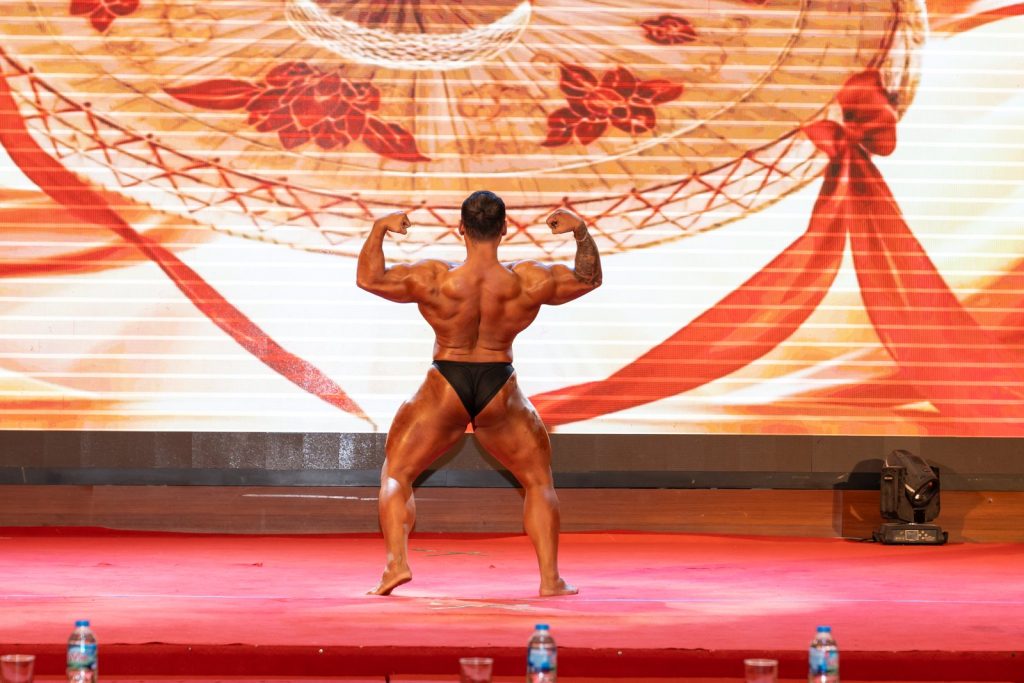 Một vận động viên Bodybuilding gồng lưng trên sân khấu có nền màn hình hình nón lá