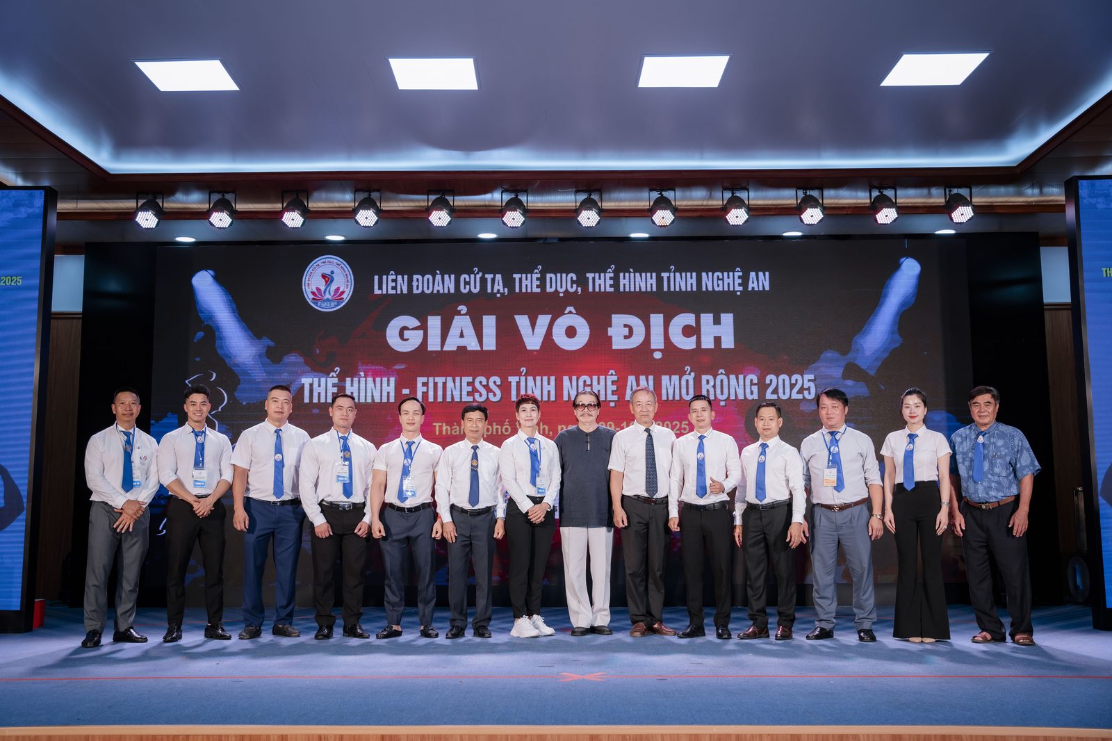 Hàng trọng tài mặc đồng phục sơ mi trắng cà vạt xanh đứng trên sân khấu Giải vô địch Thể hình – Fitness tỉnh Nghệ An mở rộng 2025