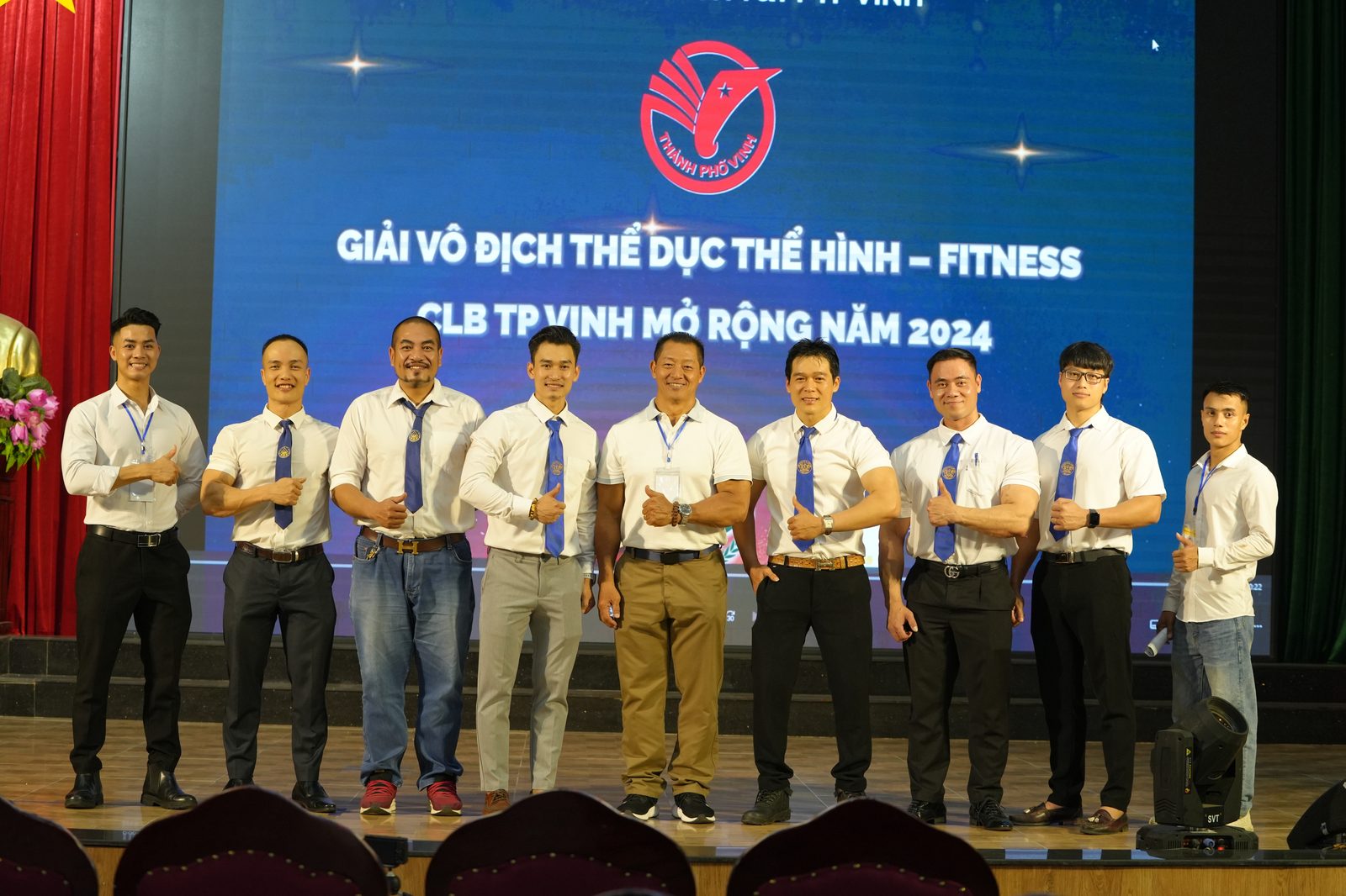 Chín người mặc đồng phục trọng tài đứng trên sân khấu Giải vô địch Thể dục Thể hình – Fitness CLB thành phố Vinh mở rộng năm 2024