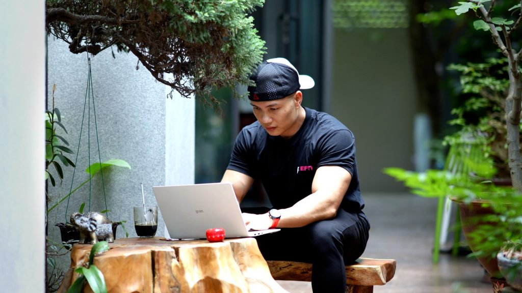 Nguyễn Bảo Bằng mặc áo thun đen ngồi làm việc với laptop trên bàn gỗ ngoài hiên, nền cây xanh