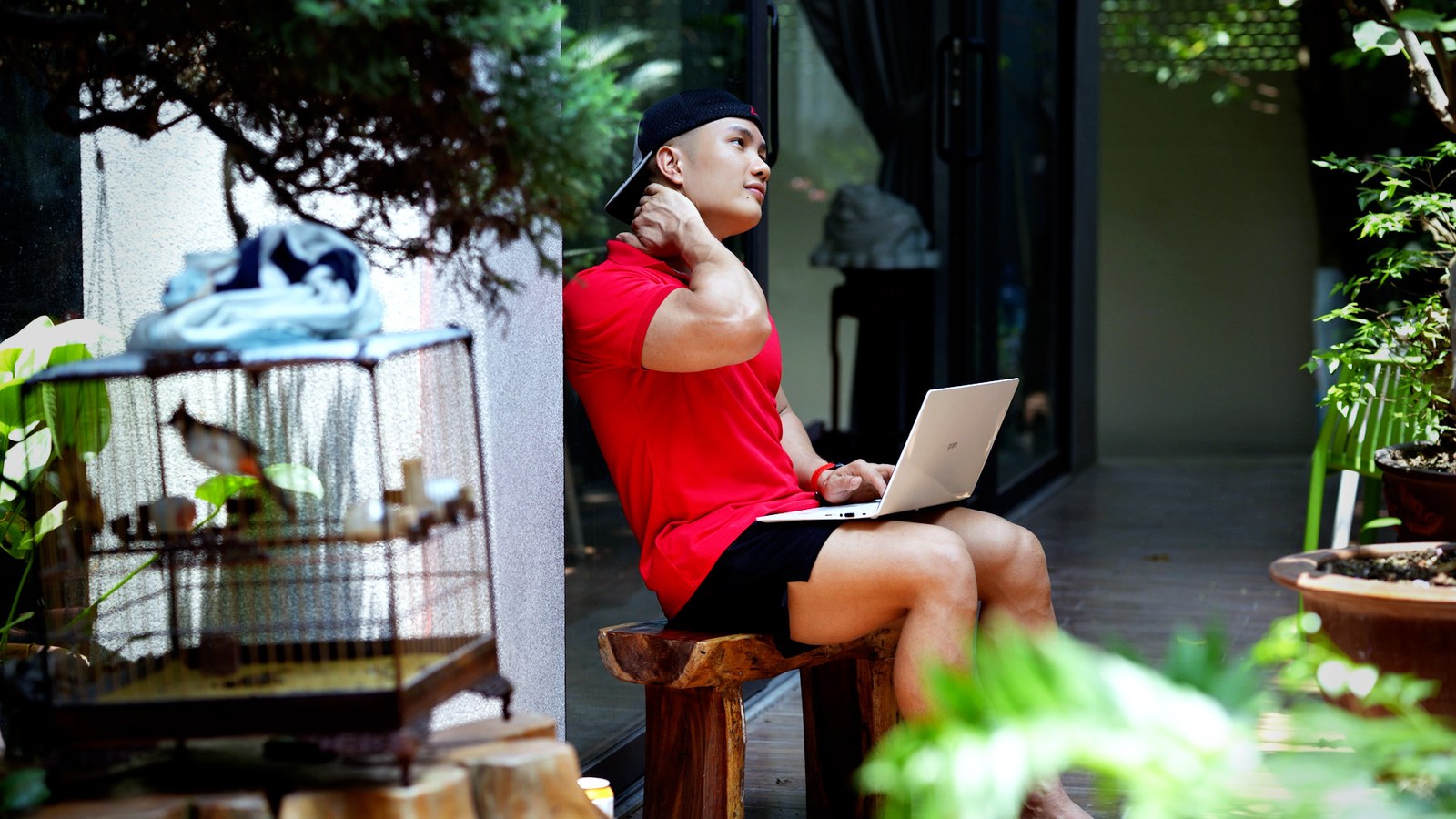 Nguyễn Bảo Bằng mặc áo thun đỏ ngồi trên ghế gỗ ngoài sân, đặt laptop trên đùi