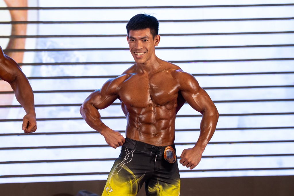 Cận một vận động viên nam cười tươi, tay chống hông, ngực bụng bóng dầu phản đèn rất rõ, quần short đen vàng
