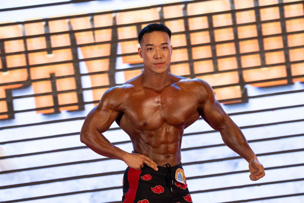 Cận một vận động viên Men Physique chống hông, da nâu đỏ bóng, múi bụng và ngực nét dưới đèn