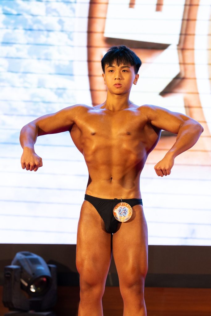 Cận nửa người một vận động viên nam gồng cơ trên sân khấu, da nâu bóng dưới đèn, nền LED trắng