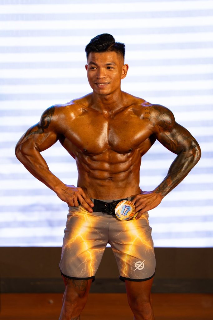 Cận cảnh một vận động viên nam Men Physique tay chống hông, da nâu đậm bóng dầu, cơ bụng và vai nét dưới đèn