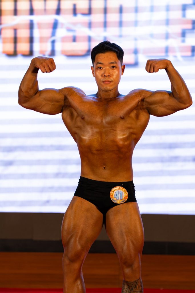 Cận một vận động viên Athletic Physique nam gồng hai tay trước, màu da nâu cam bóng dưới đèn