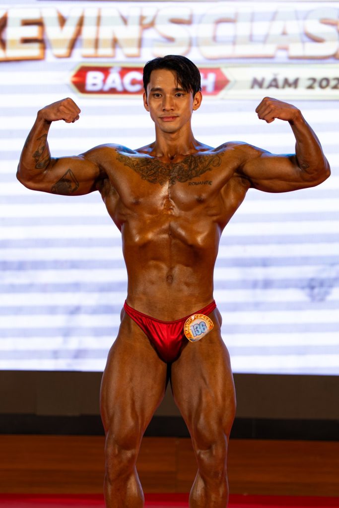 Cận một vận động viên bodybuilding nam quần đỏ gồng hai tay trước, màu tan nâu đỏ bóng, nền LED