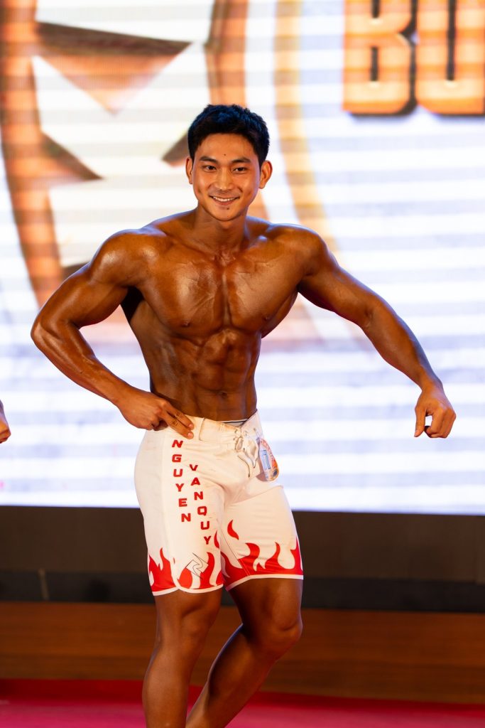 Một vận động viên Men Physique quần trắng chống hông, da nâu đậm bóng dầu, ảnh dọc cận 3/4 người