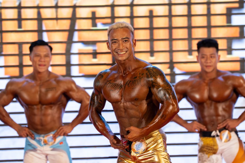 Ba vận động viên Men Physique cận nửa người, người giữa tóc bạch kim màu da ánh vàng đồng, hai bên nâu đỏ, tương phản tông màu rõ
