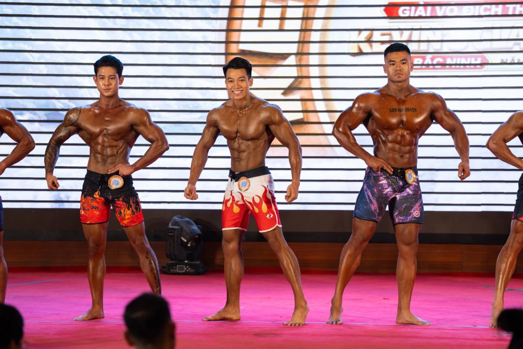 Nhóm vận động viên Men Physique xếp hàng ngang tầm, tông màu da khác nhau từ nâu đỏ tới nâu sẫm, quần short nhiều màu