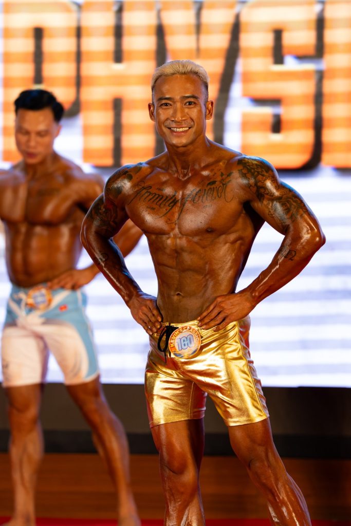 Hai vận động viên Men Physique đứng cạnh nhau, người quần vàng kim da đậm hơn hẳn người bên trái, thấy chênh lệch màu tan