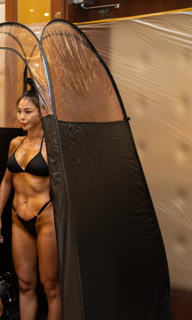 Một vận động viên nữ bikini đen đứng trong lều phun, da nâu bóng sau khi phun màu