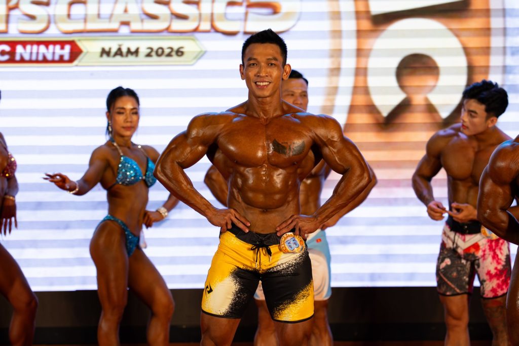 Vòng toàn năng: vận động viên nữ bikini xanh đứng cạnh vận động viên nam quần vàng đang gồng, da bóng dưới đèn