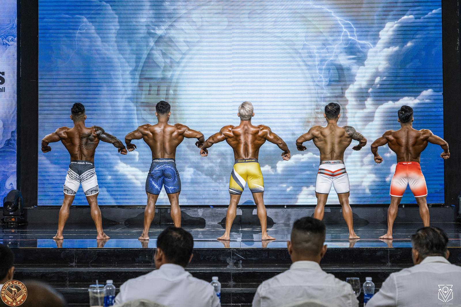 Hàng vận động viên Men Physique quay lưng, trọng tài ngồi chấm