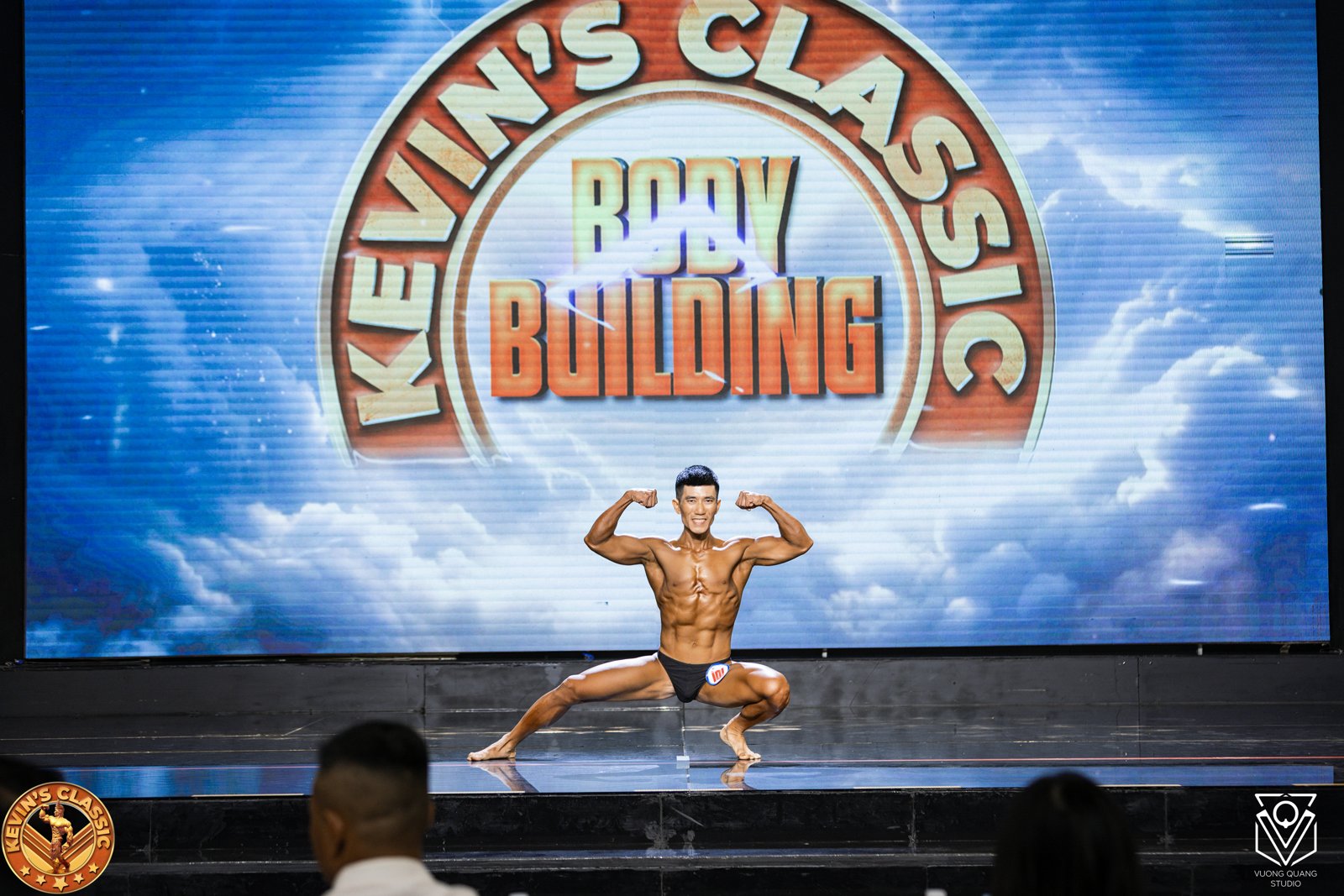 Một vận động viên tạo dáng giữa sân khấu rộng trước phông Body Building