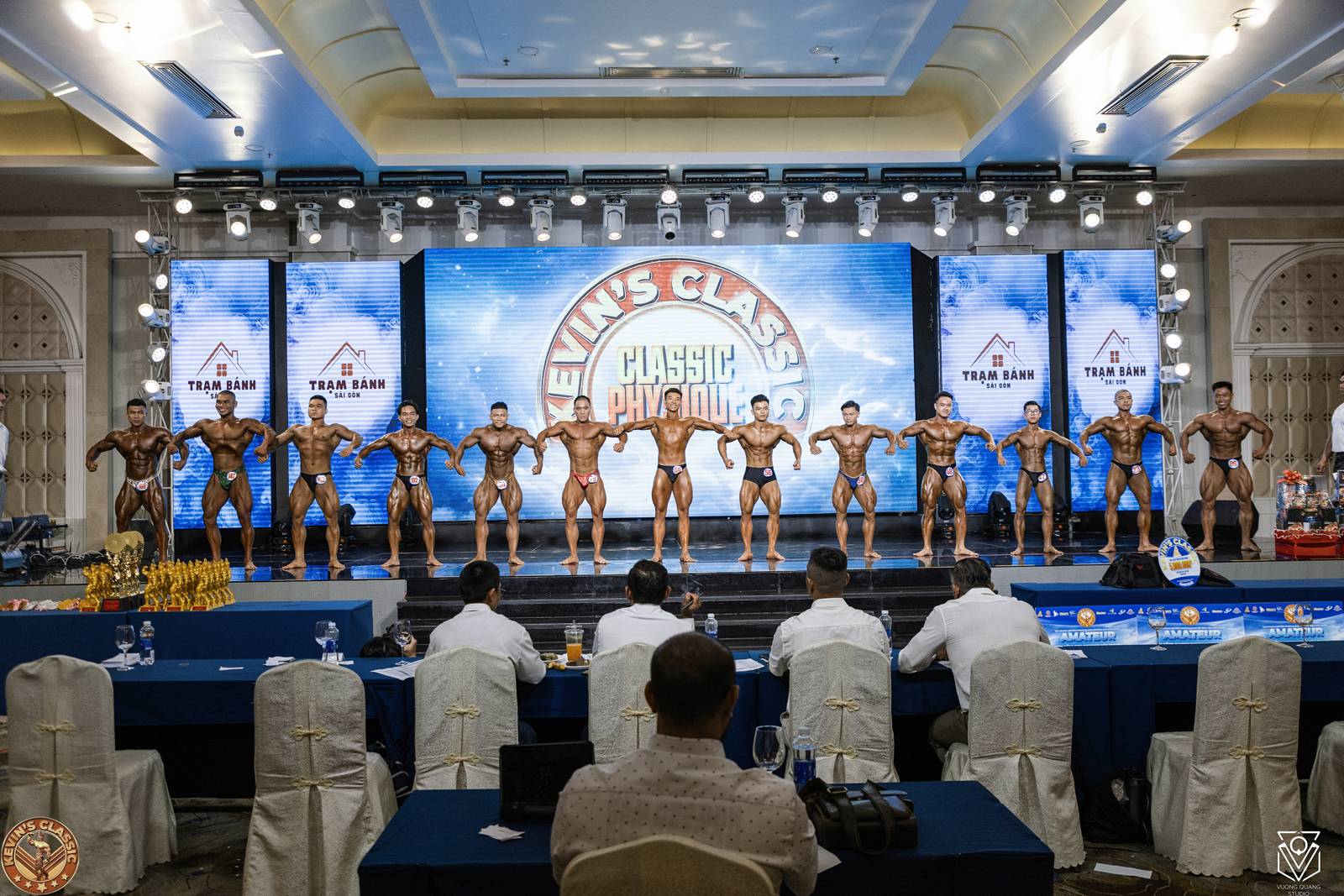 Hàng vận động viên Classic Physique trên sân khấu, trọng tài ngồi chấm phía trước
