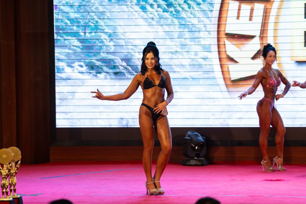 Một vận động viên bikini bộ đen dang tay tạo dáng ở tiền cảnh, một vận động viên khác phía sau, nền LED