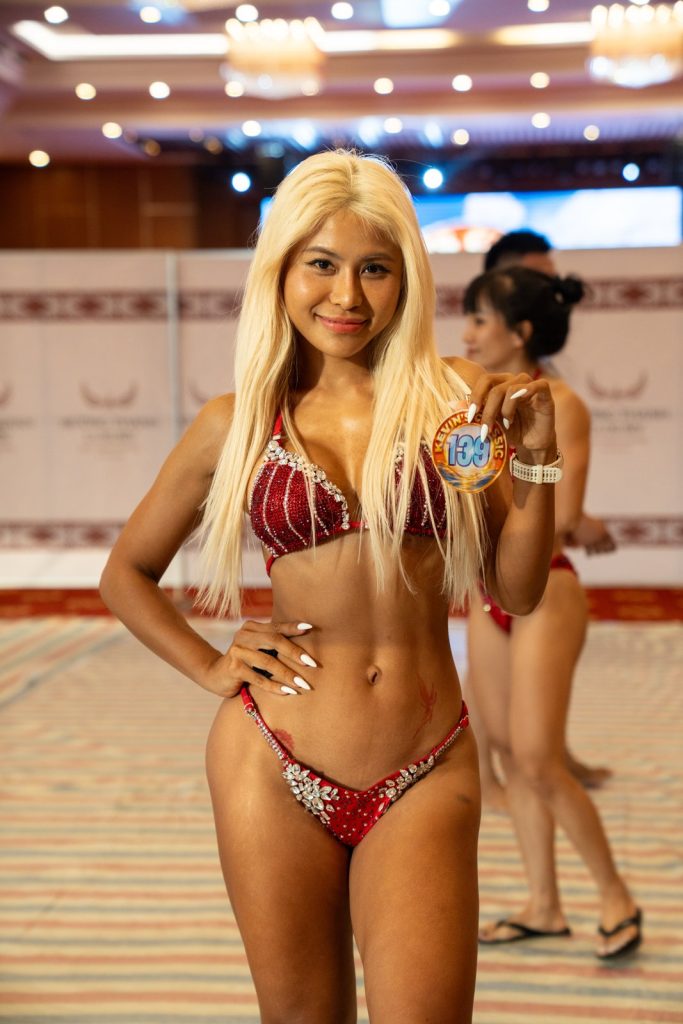 Một vận động viên tóc vàng bikini đỏ chụp chân dung nửa người ở hậu trường, da đã tan màu, tay chống hông