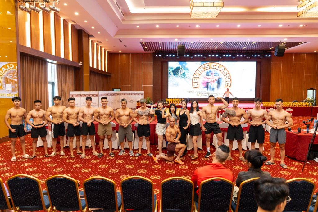 Nhóm vận động viên đứng tạo dáng trước sân khấu trong hội trường ngày cân đo