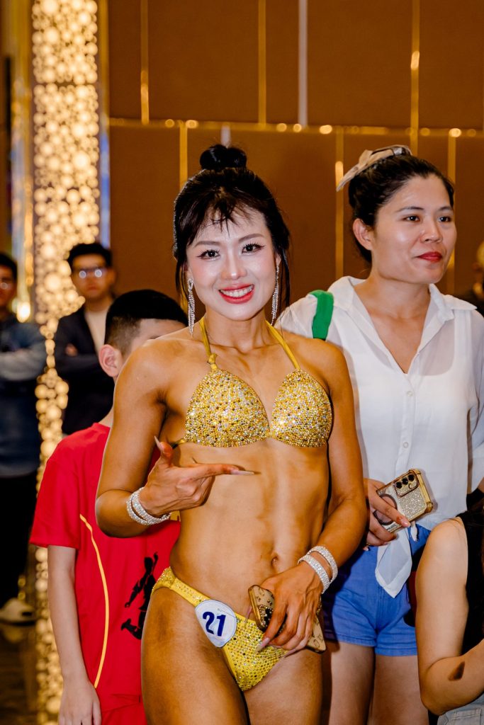 Hậu trường, vận động viên nữ cười cùng người hỗ trợ tại giải thể hình Nghệ An 2026
