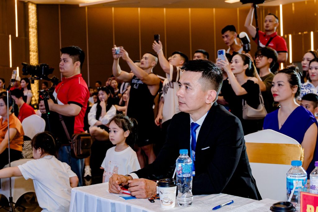 Ông Nguyễn Song Hào, Chủ tịch Liên đoàn Cử tạ, Thể dục, Thể hình tỉnh Nghệ An, ngồi bàn đại biểu xem thi, khán giả phía sau – giải thể hình Nghệ An 2026