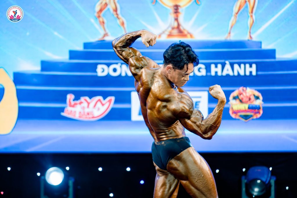 Cận cảnh một vận động viên thể hình xoay lưng gồng tay trước dòng Đơn vị đồng hành tại giải thể hình Nghệ An 2026