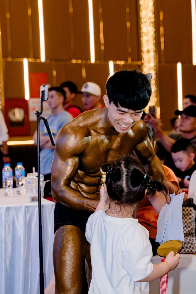 Vận động viên đeo huy chương cúi xuống với một bé gái tại giải thể hình Nghệ An 2026