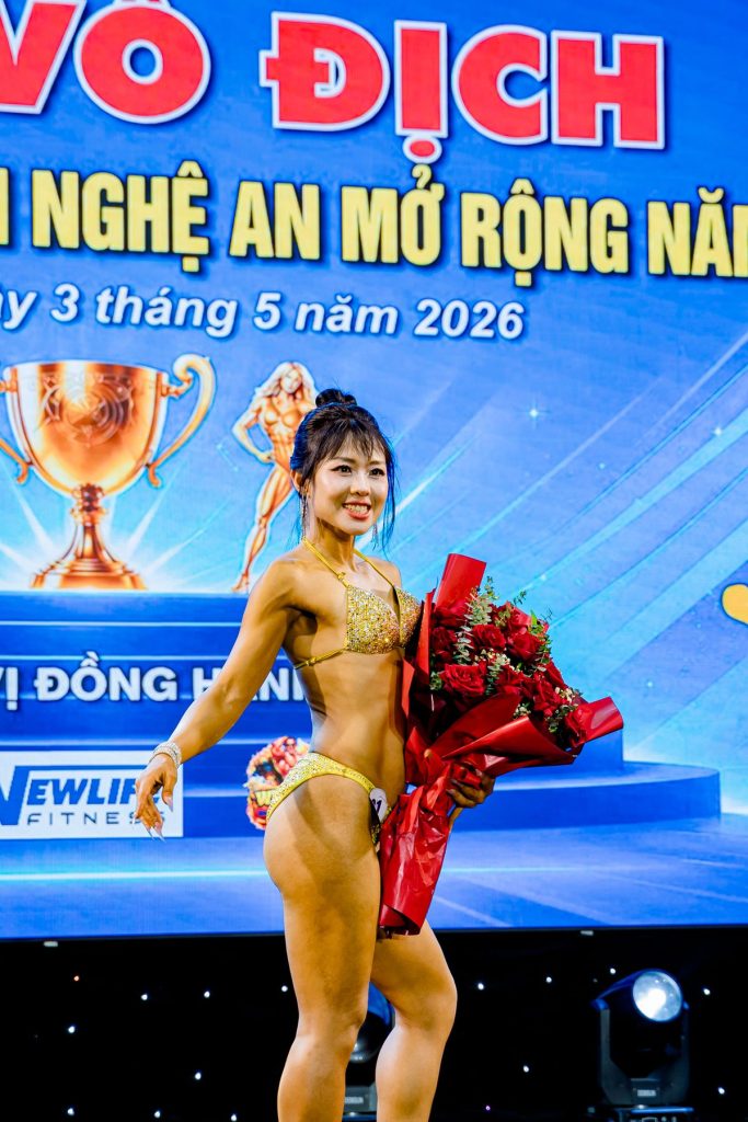 Vận động viên nữ ôm bó hoa cười trên sân khấu tại giải thể hình Nghệ An 2026