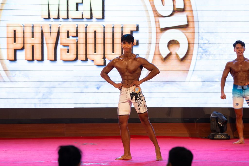 Vận động viên trẻ quần short trắng tạo dáng mặt trước, phía sau là phông chữ Men Physique
