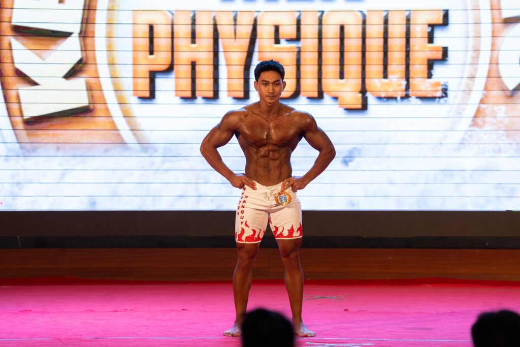 Vận động viên trẻ quần short trắng họa tiết đỏ tạo dáng mặt trước, phía sau là chữ Physique