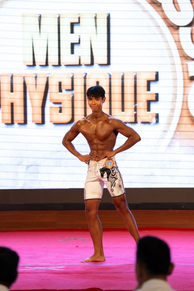 Vận động viên trẻ quần short trắng tạo dáng mặt trước, phía sau là phông chữ Men Physique