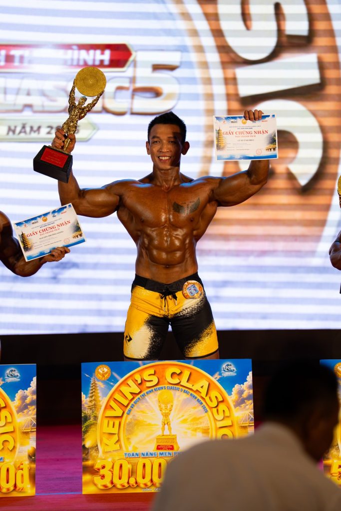 Vận động viên giơ cúp và giấy chứng nhận, phía trước là bảng thưởng toàn năng Men Physique