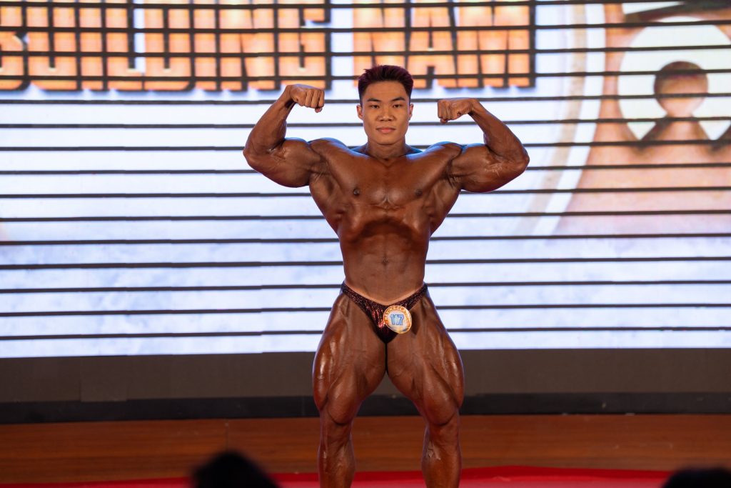 Vận động viên gồng hai tay trước, phía sau là phông chữ Bodybuilding Nam