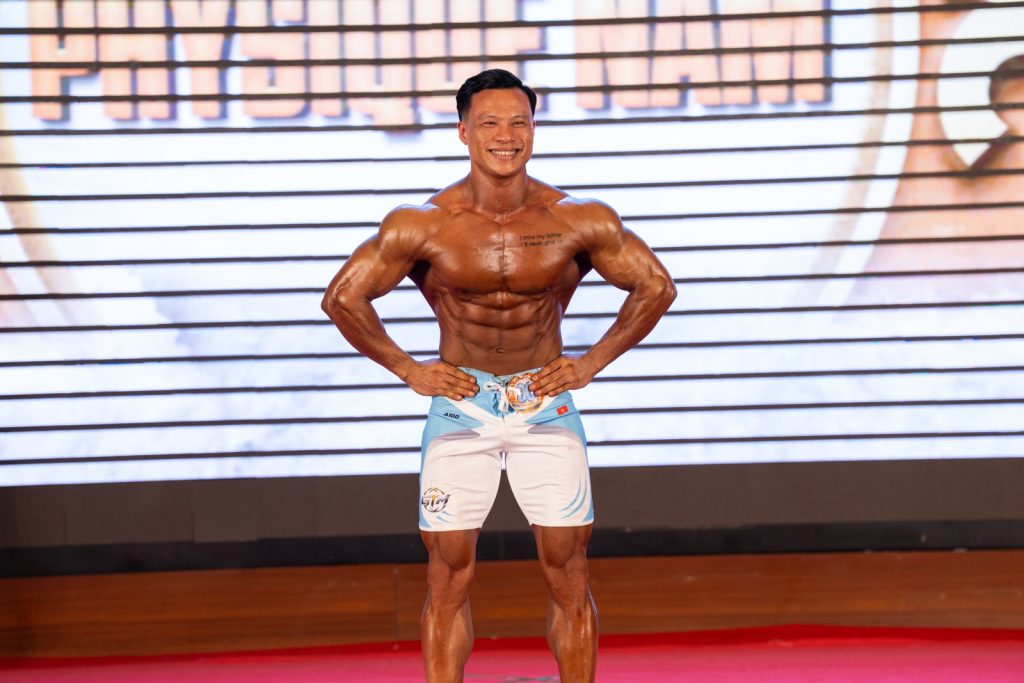 Vận động viên quần short trắng xanh tạo dáng mặt trước, hai tay chống hông, trước phông chữ Physique Nam