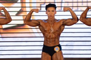 Vận động viên gồng hai tay trước, cùi chỏ ngang vai, nhìn thẳng, cận nửa người trên sân khấu