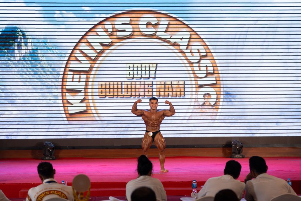 Vận động viên gồng hai tay trước trên sân khấu, hàng ghế phía dưới nhìn lên